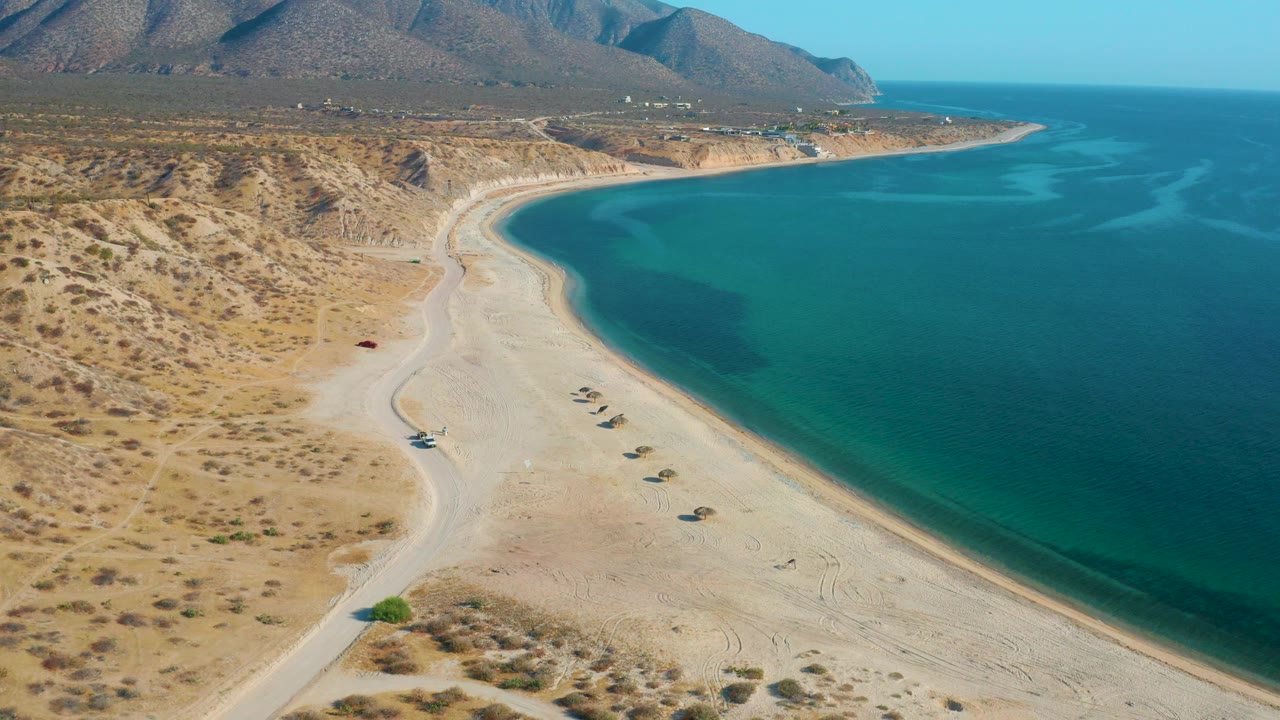Mid-clip aerial frame over El Sargento La Ventana showing desert scrub in the lower left, pale sand along the coast, blue water to the right, and no visible people.