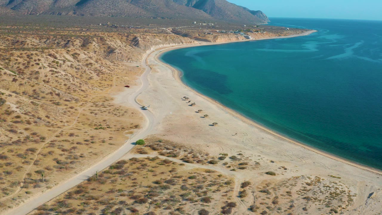 Opening aerial frame over El Sargento La Ventana, Mexico, with scrubby desert foreground, a pale beach curving along blue water, low mountains beyond, and no visible people.