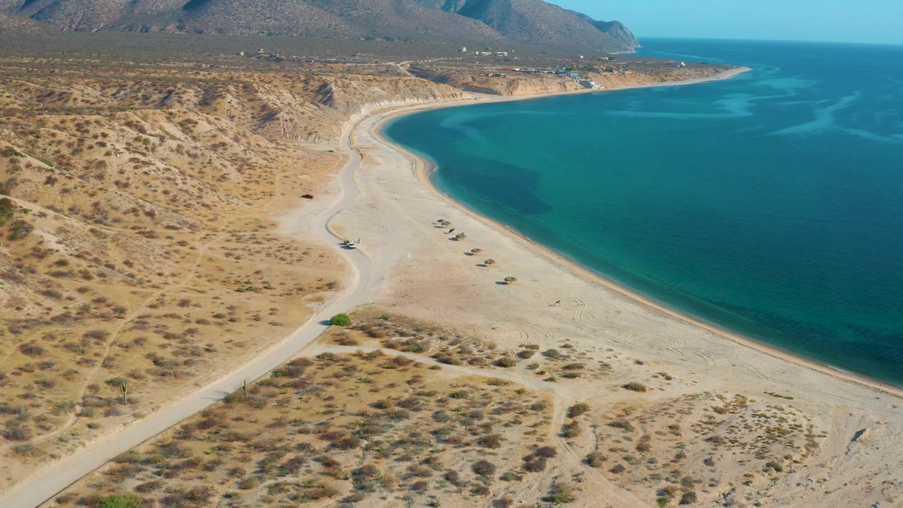 The aerial angle over El Sargento La Ventana, Mexico, holds a broad view of pale sand, blue bay water, scrub-covered ground, distant mountains, and no visible people.