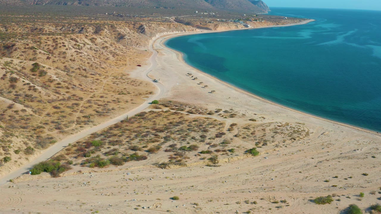 Aerial view in El Sargento La Ventana, Mexico, with a dirt road cutting through tan desert scrub toward the beach and bright blue water, with no visible people.