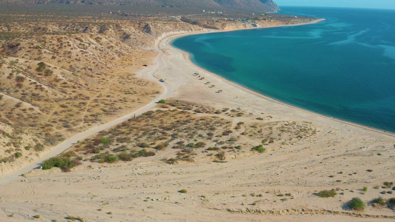 Drone-style aerial frame over El Sargento La Ventana, Mexico, showing the beach curve, low brush on sandy ground, turquoise water on the right, and no visible people.