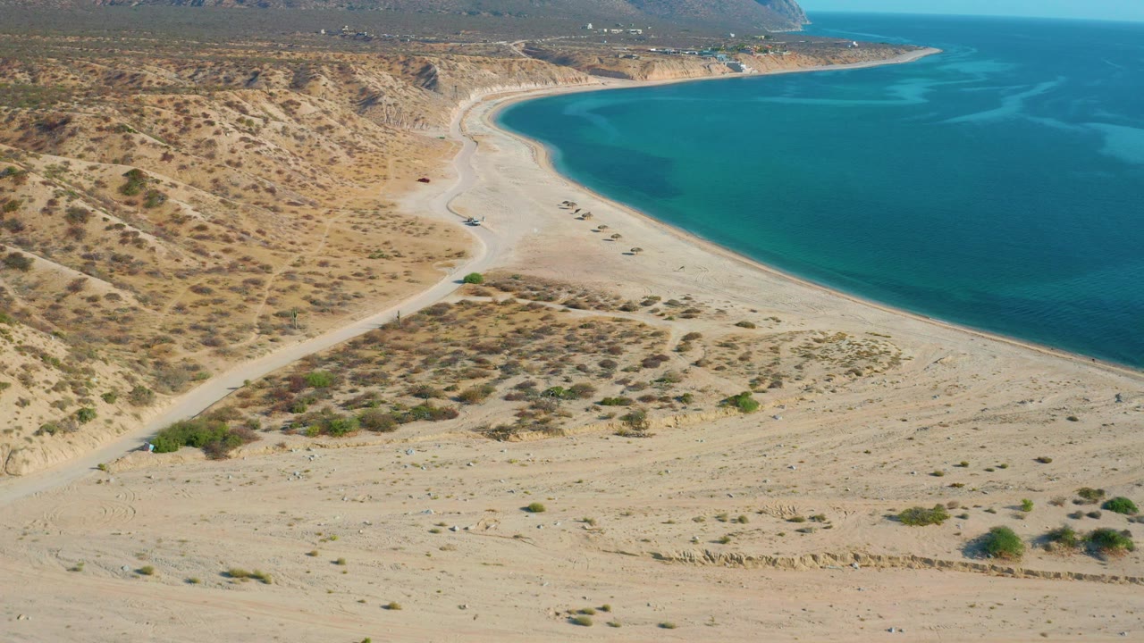 Aerial view of El Sargento La Ventana, Mexico, with a wide sandy foreground, desert hillside to the left, calm blue water to the right, and no visible people.