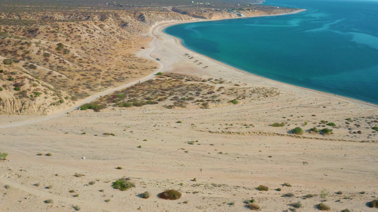 High view in El Sargento La Ventana, Mexico, showing low desert brush across the sandy foreground, a pale beach curve, blue water, and no visible people.