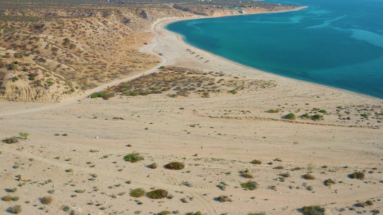 A mid-clip aerial frame in El Sargento La Ventana, Mexico, with faint vehicle tracks through sand, small shrubs, tan bluffs, blue bay water, and no visible people.