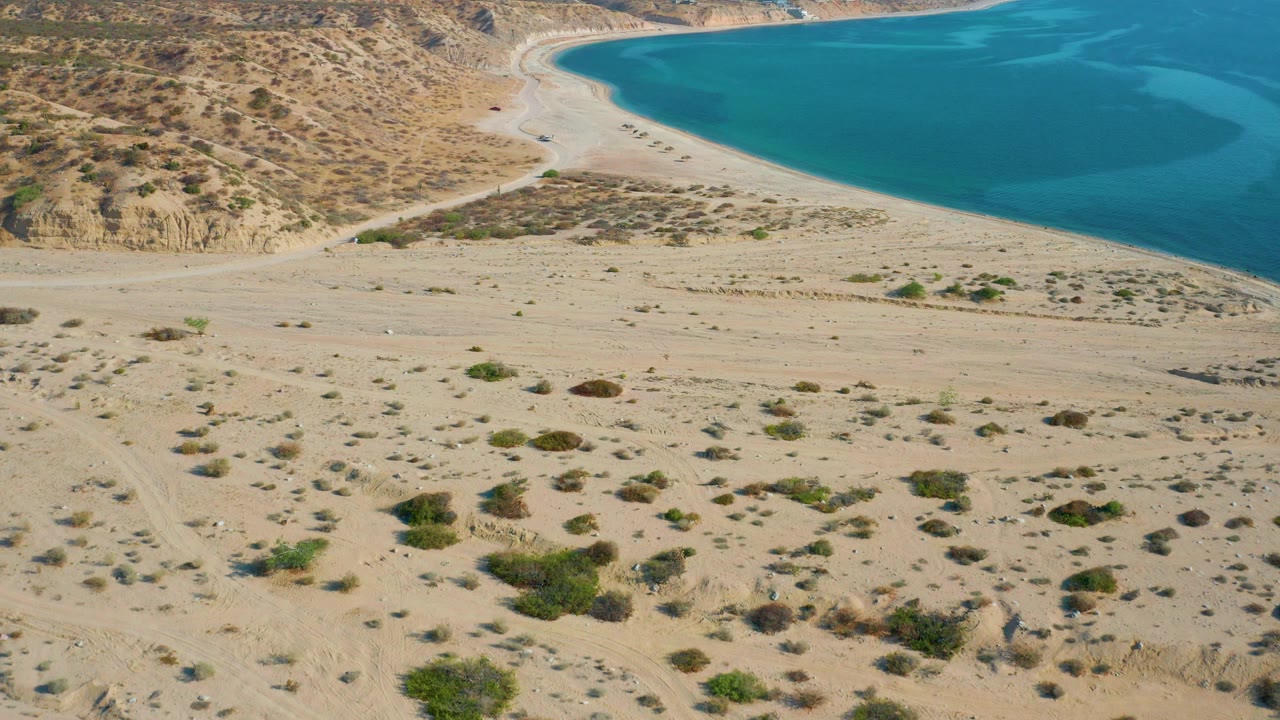Aerial frame over El Sargento La Ventana showing pale sand, scrub patches, tire tracks, a curved turquoise bay, dry hills, and no visible people.