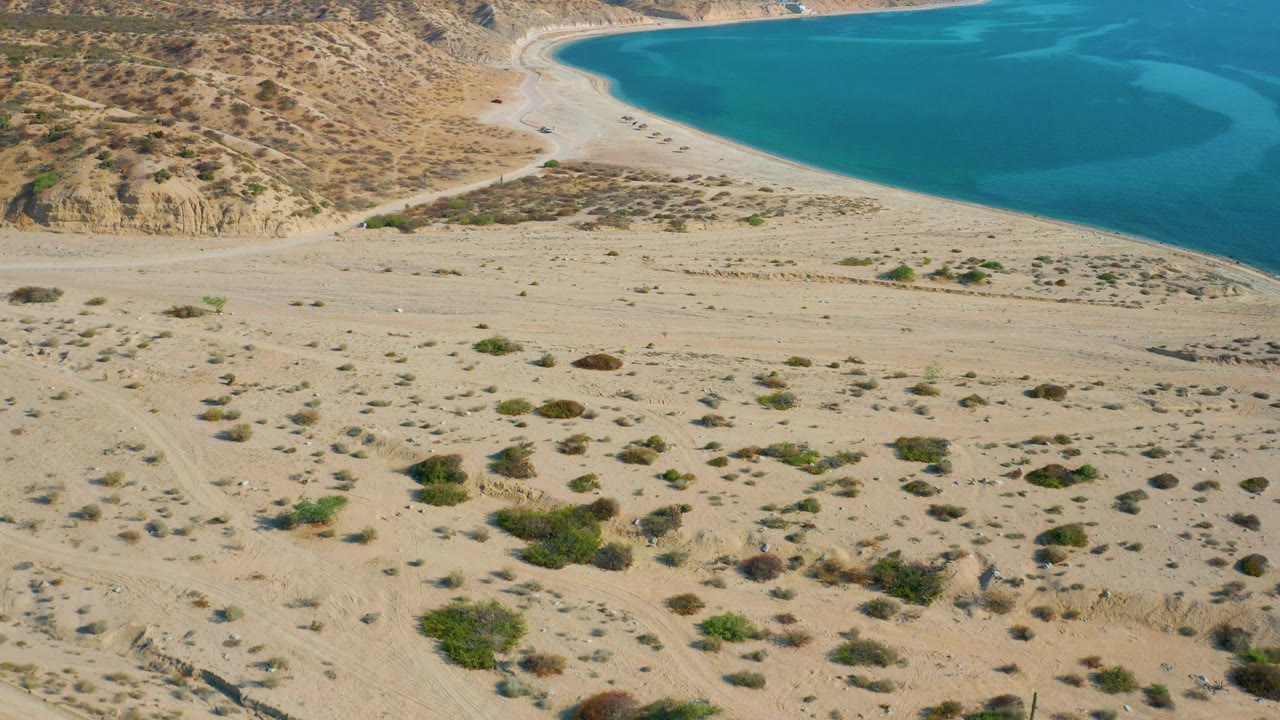 High aerial frame over El Sargento La Ventana showing desert scrub and sandy tracks leading toward a pale crescent beach and turquoise water, with no visible people.