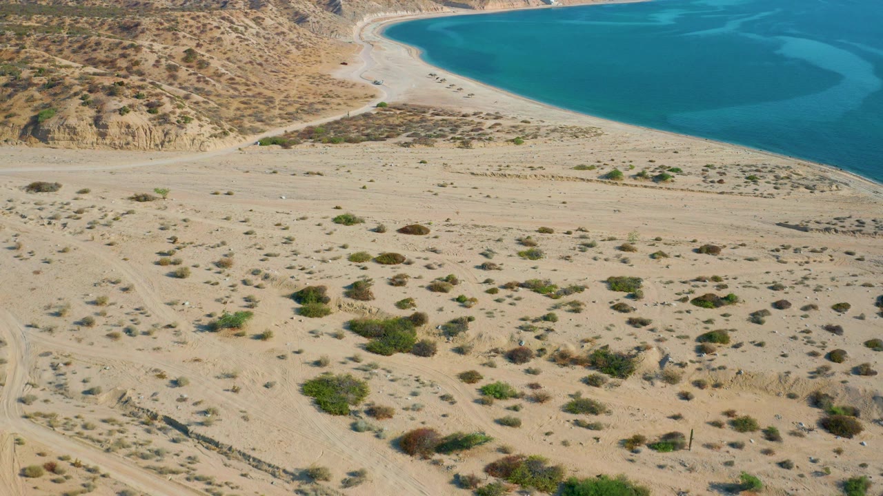 Wide aerial of empty beach below dry hills and blue sea.