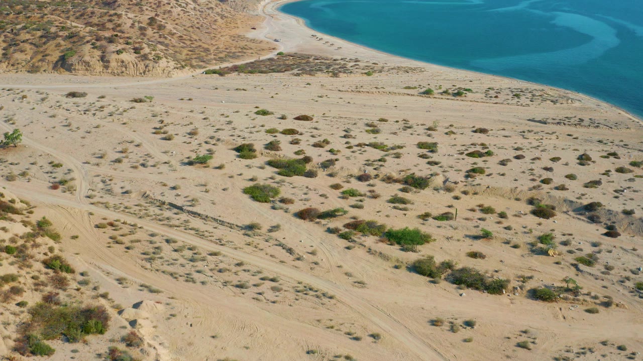Aerial frame in El Sargento La Ventana, Mexico showing sandy scrubland, dirt tracks, a pale beach curve, and blue coastal water at the top, with no visible people.