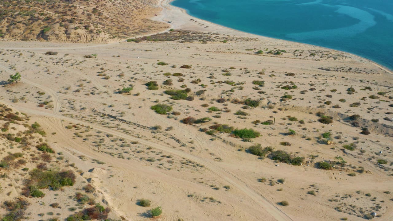 Aerial frame in El Sargento La Ventana, Mexico showing a wider spread of scrub patches across sand, a curving beach line, and turquoise water in the background, with no visible people.