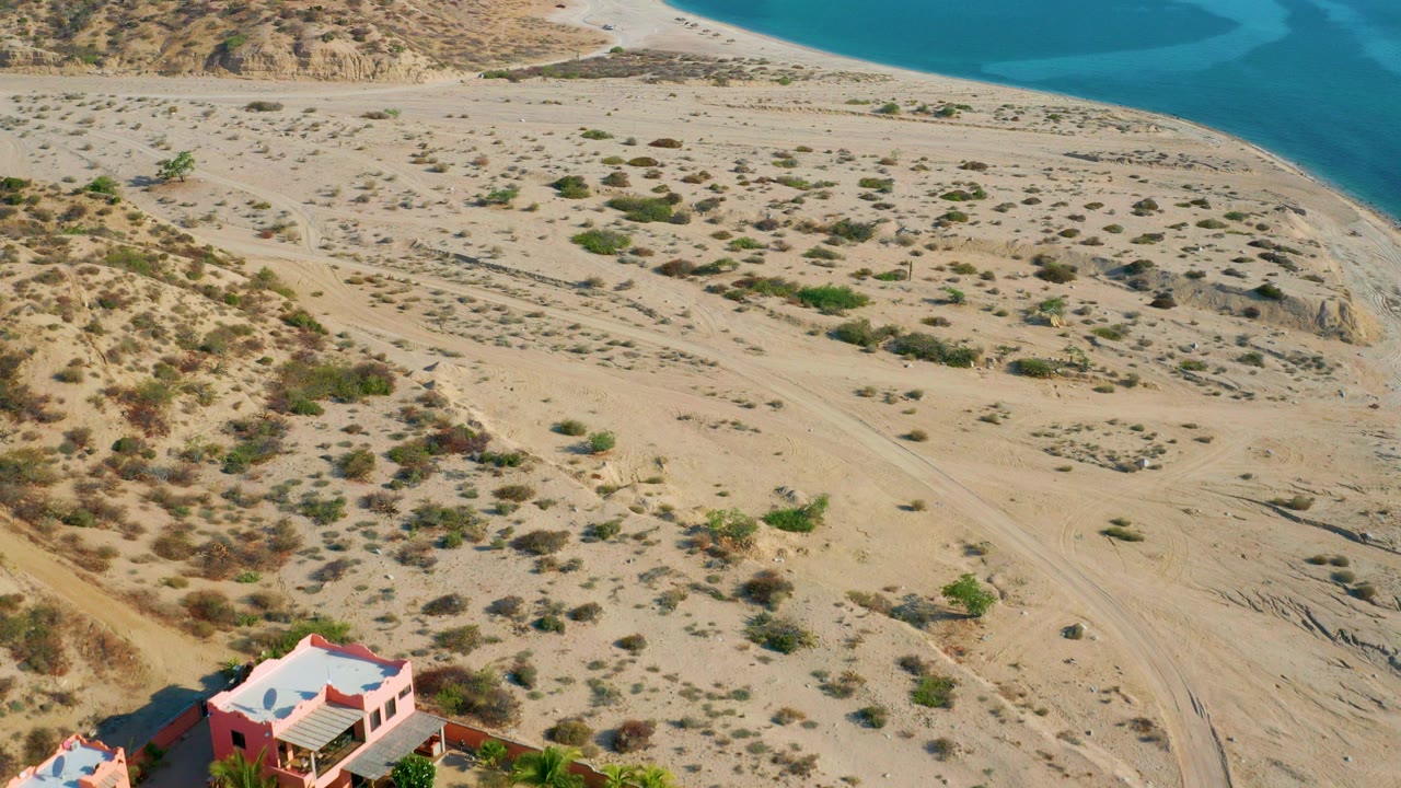 Aerial frame from El Sargento La Ventana, Mexico showing sandy desert terrain, blue coastal water, and the edge of a coral-colored home compound with no visible people.