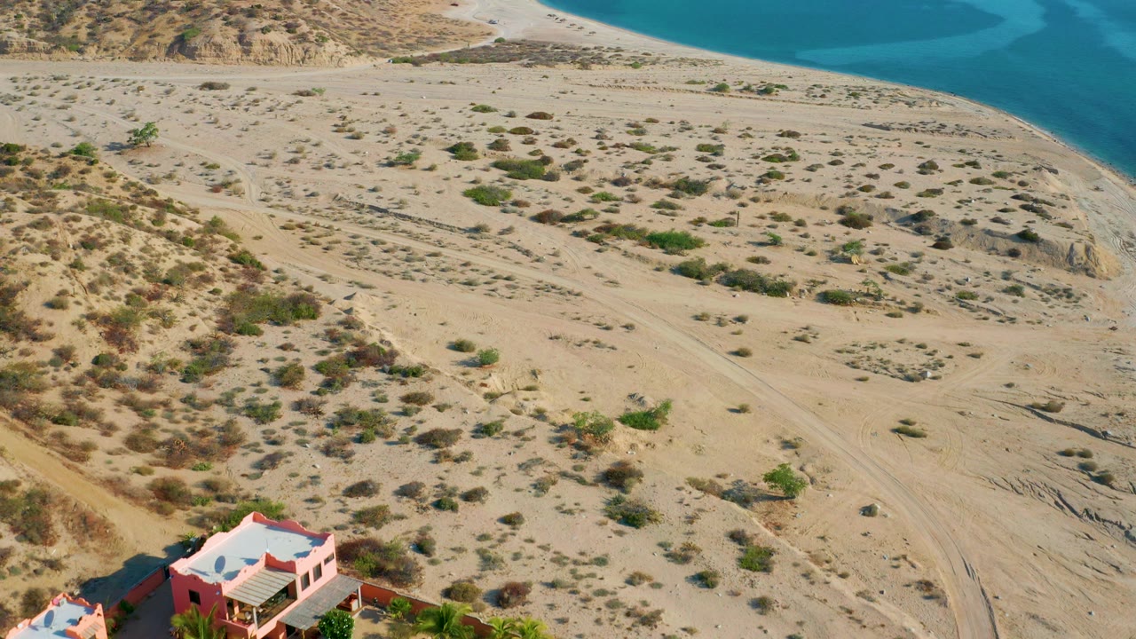 Aerial view in El Sargento La Ventana, Mexico with a coral-colored home partly visible at the bottom, scrub-covered sand across the middle, and blue sea beyond, with no visible people.