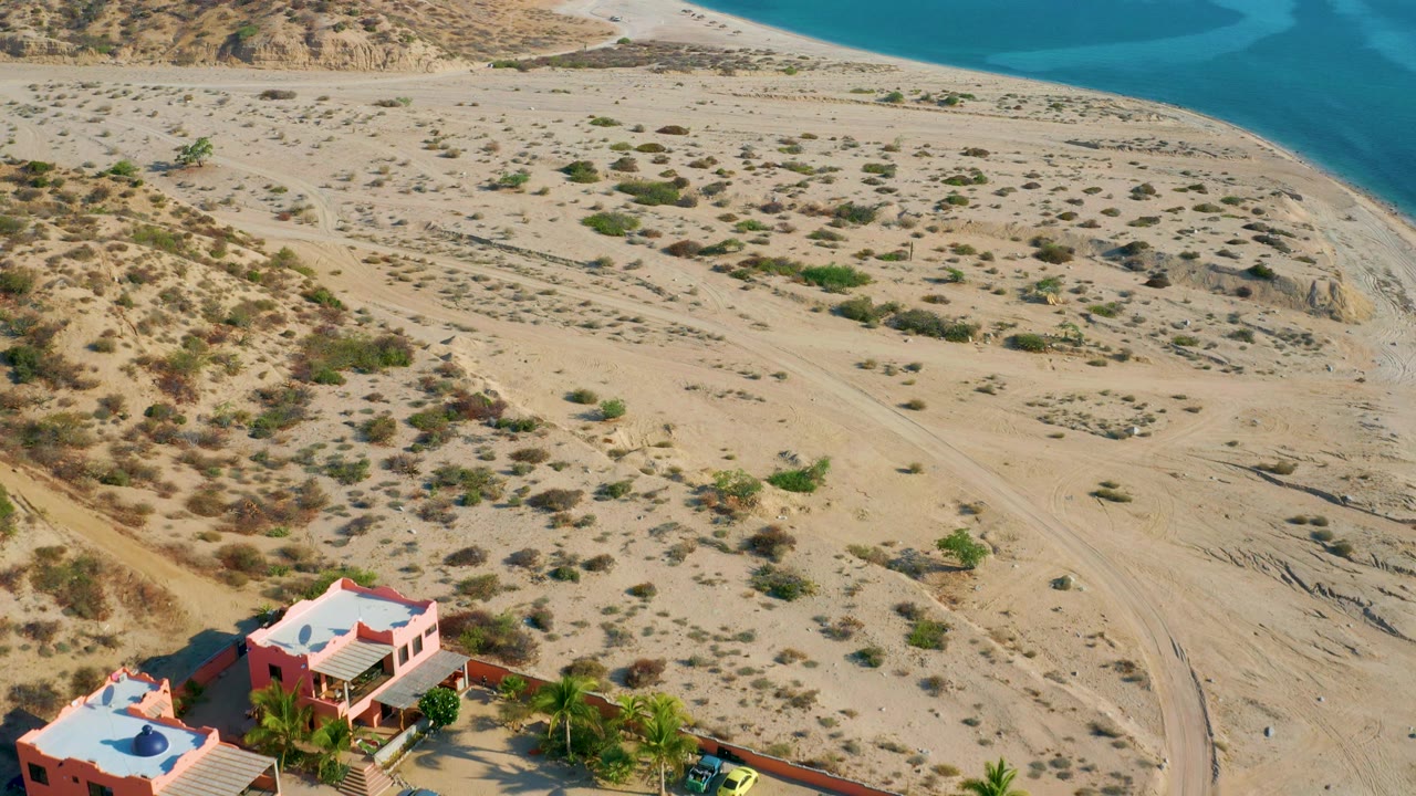 Drone perspective over El Sargento La Ventana, Mexico showing more of the coral-walled home compound, palm trees, sandy beach flats, and blue water in the background, with no visible people.