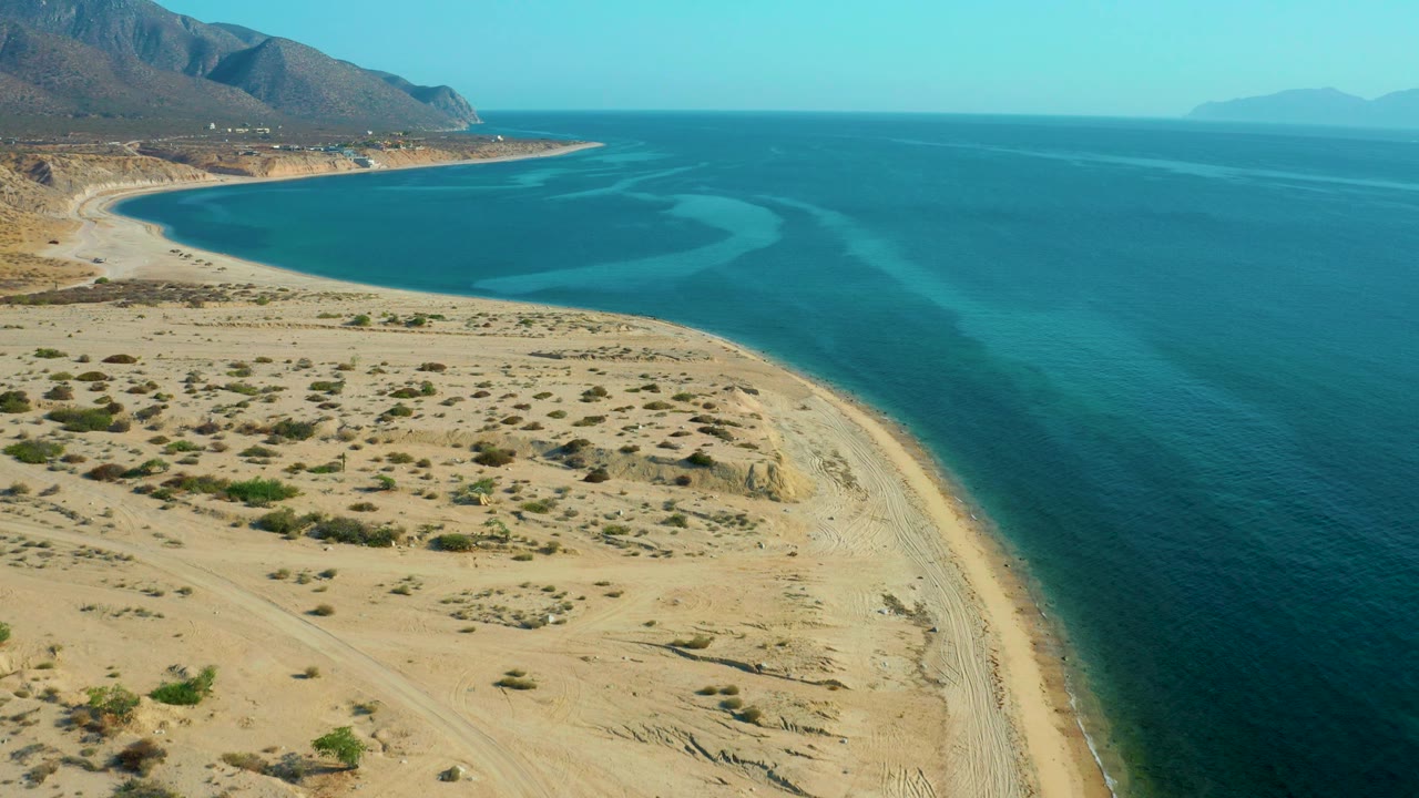 Elevated view of the El Sargento La Ventana coast with scrub-covered sand, a narrow beach edge, turquoise shallows, darker offshore water, and no visible people.