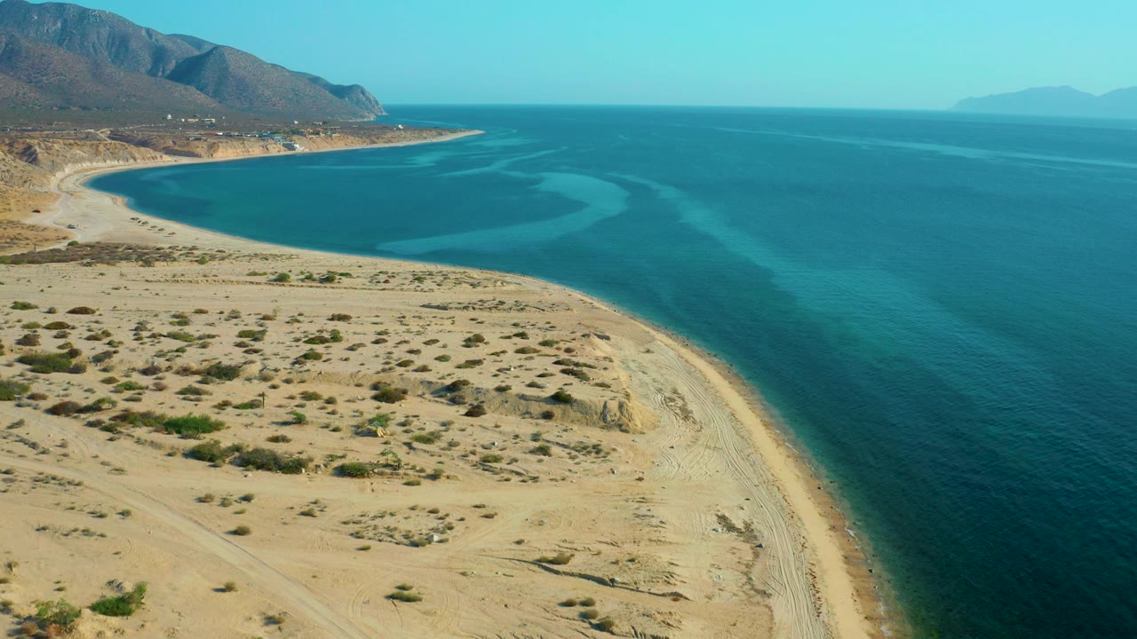 Aerial view in El Sargento La Ventana, Mexico, showing the sandy desert foreground, the beach wrapping around a blue bay, hazy distant landforms, and no visible people.
