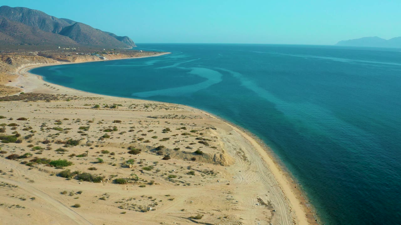 Wide aerial frame of the El Sargento La Ventana shoreline, with textured sand and sparse shrubs below, calm blue sea across the right side, and no visible people.