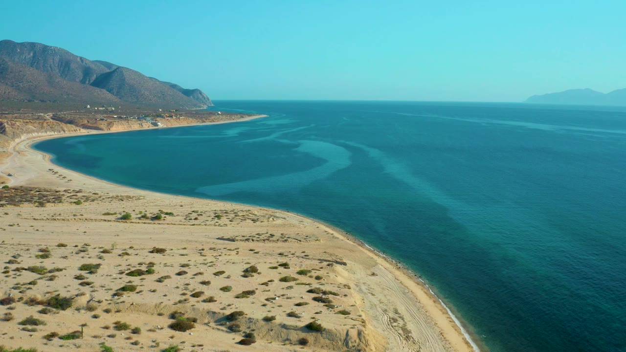 At 0:01, the aerial composition holds over El Sargento La Ventana with desert scrub in the foreground, a rounded beach line, layered blue water, mountains at left, and no visible people.