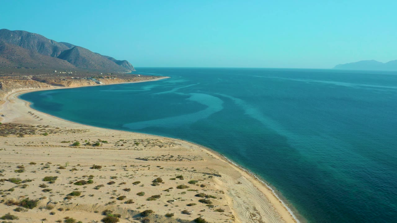 At 0:02, the frame shows the empty sand and scrub of El Sargento La Ventana below a smooth turquoise bay, with a mountain ridge on the left and open blue sea to the right, no visible people.
