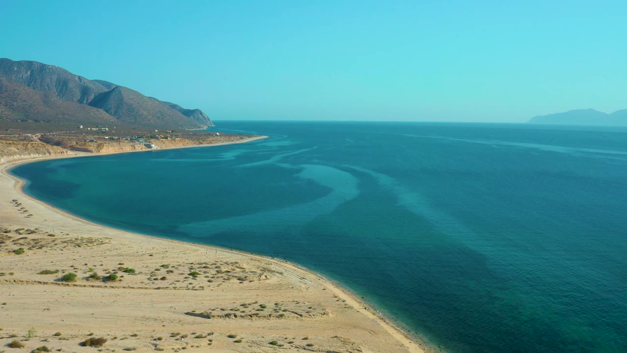 Drone-height coastal view in El Sargento La Ventana, Mexico, showing pale beach sand below, blue water bands through the bay, distant mountains, and no visible people.