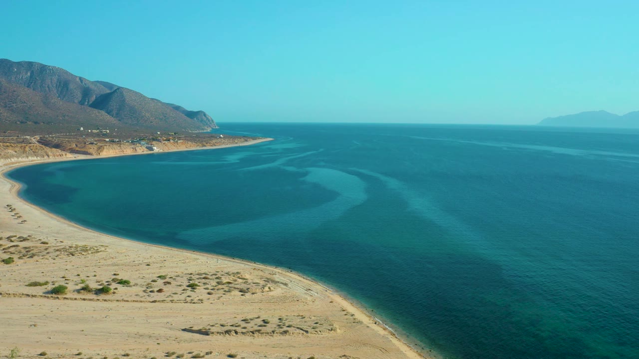 High view over El Sargento La Ventana, Mexico, with the curved beach low in frame, shallow turquoise water tracing the bay, open sea to the right, and no visible people.