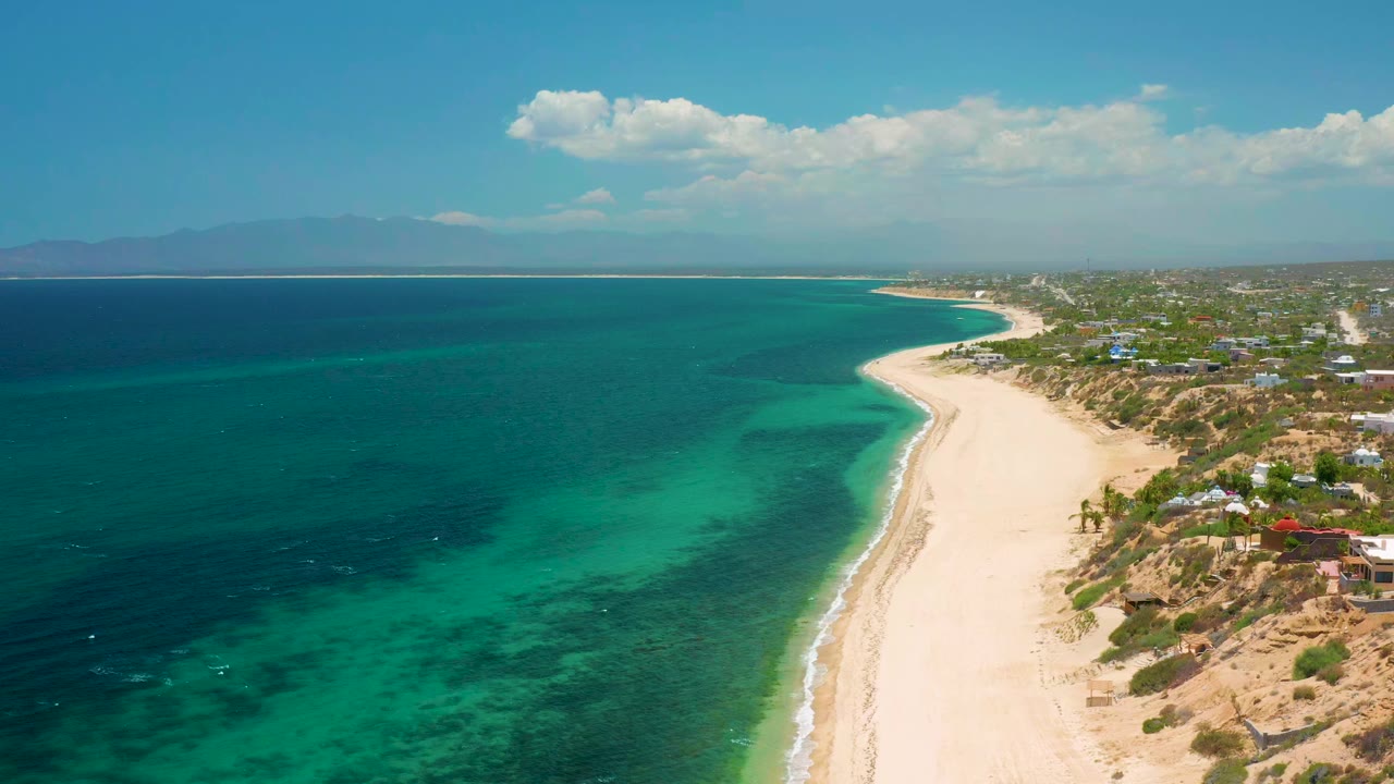 Opening aerial frame over El Sargento La Ventana, Mexico, showing clear turquoise water, a pale curving beach, inland greenery and buildings, distant mountains, and no visible people.