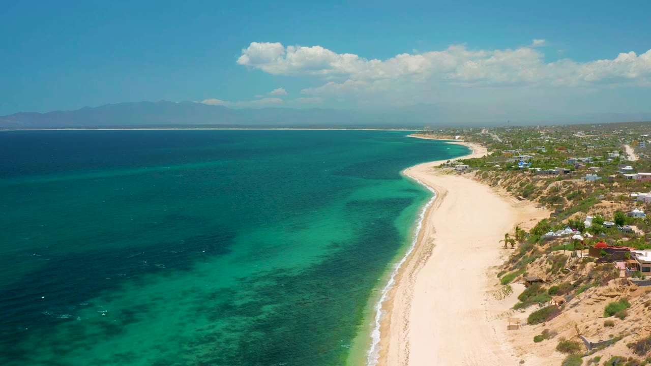 Aerial frame in El Sargento La Ventana, Mexico, with the beach curve more centered, dark and light water patches offshore, low coastal buildings inland, distant mountains, and no visible people.