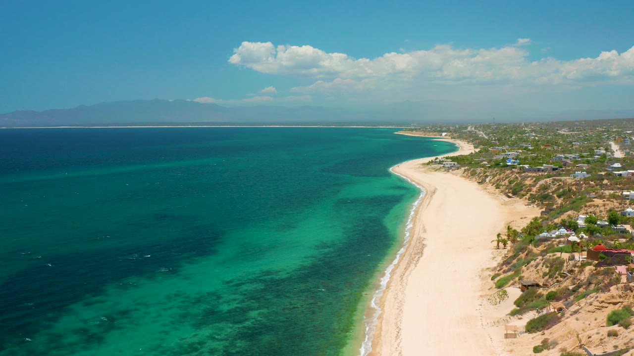 Opening aerial frame over El Sargento La Ventana, Mexico, showing turquoise shallows, a pale beach curving right, distant mountains, scattered coastal homes, and no visible people.