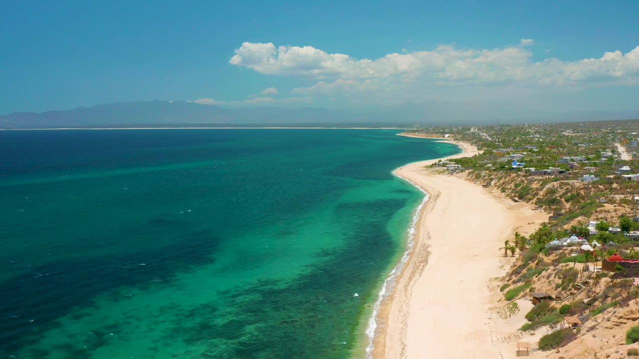 Aerial coastal view with darker offshore water, pale sand, and a small beachside settlement.