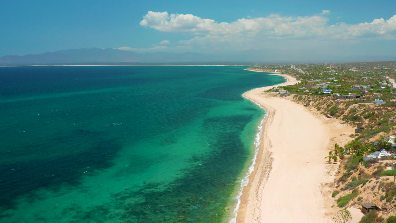 Opening aerial frame over El Sargento La Ventana, Mexico, with golden sand curving beside emerald shallows, darker offshore water, coastal foliage and buildings to the right, hazy mountains on the horizon, and no visible people.