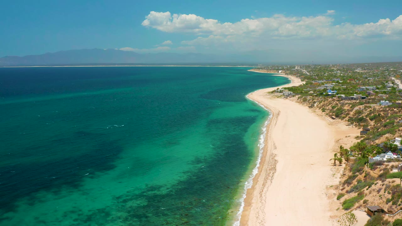 At 0:01, the aerial view holds on El Sargento La Ventana, Mexico, showing a broad band of turquoise water beside pale sand, dry shoreline vegetation, scattered low buildings, distant mountains, and no visible people.