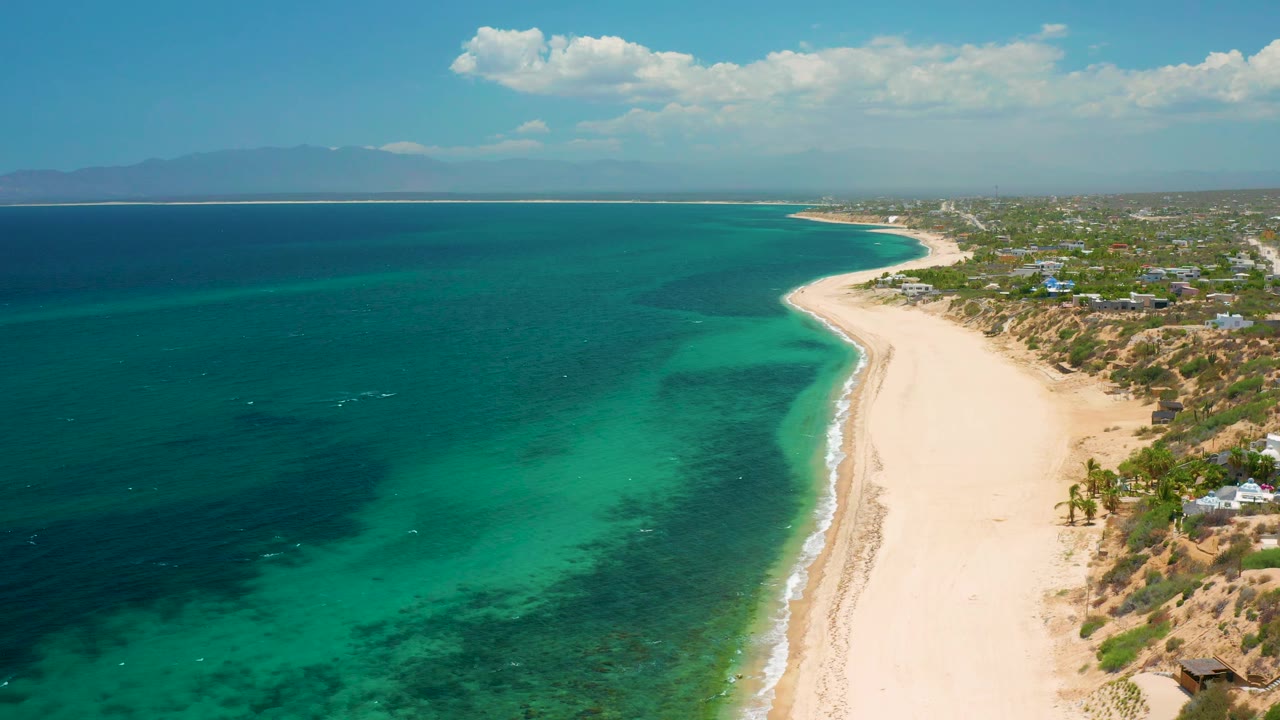 At 0:02, the frame shows El Sargento La Ventana, Mexico from above, with empty golden sand running along green-blue water, darker sea farther left, coastal homes and trees at right, hazy mountains beyond, and no visible people.