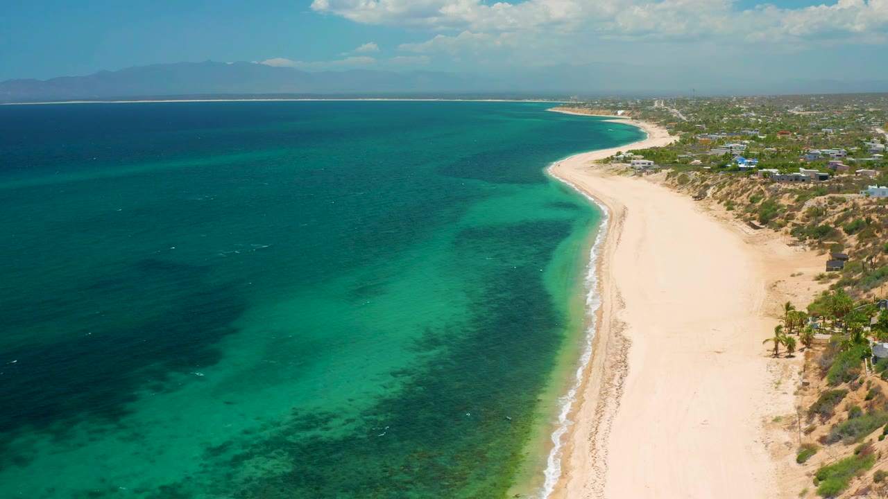 Drone-height view in El Sargento La Ventana, Mexico, with a long pale beach running up the right side, turquoise shallows, dark sea patches, a distant mountain horizon, and no visible people.