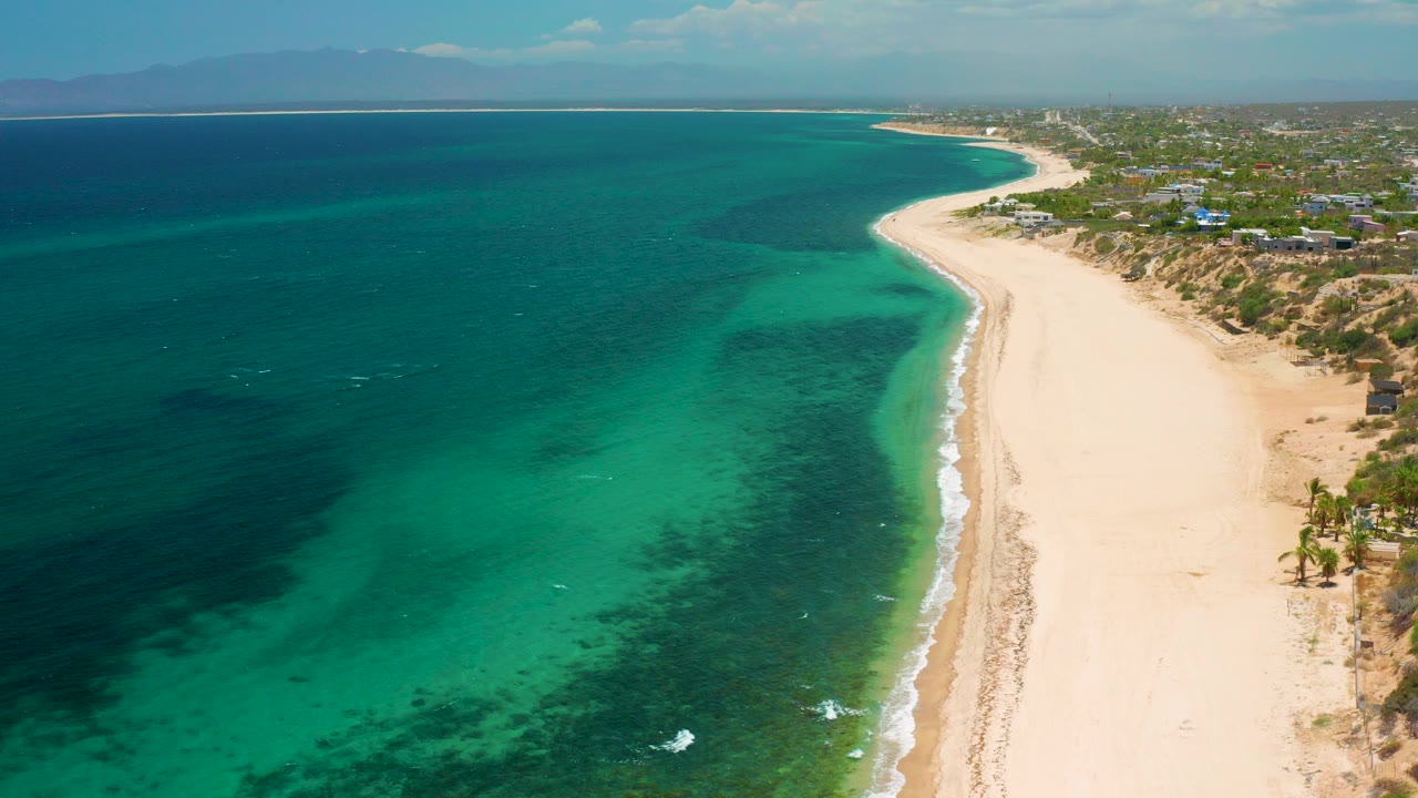 Aerial view over El Sargento La Ventana, Mexico, with turquoise water on the left, a pale empty beach on the right, seaweed near the surf line, and no visible people.