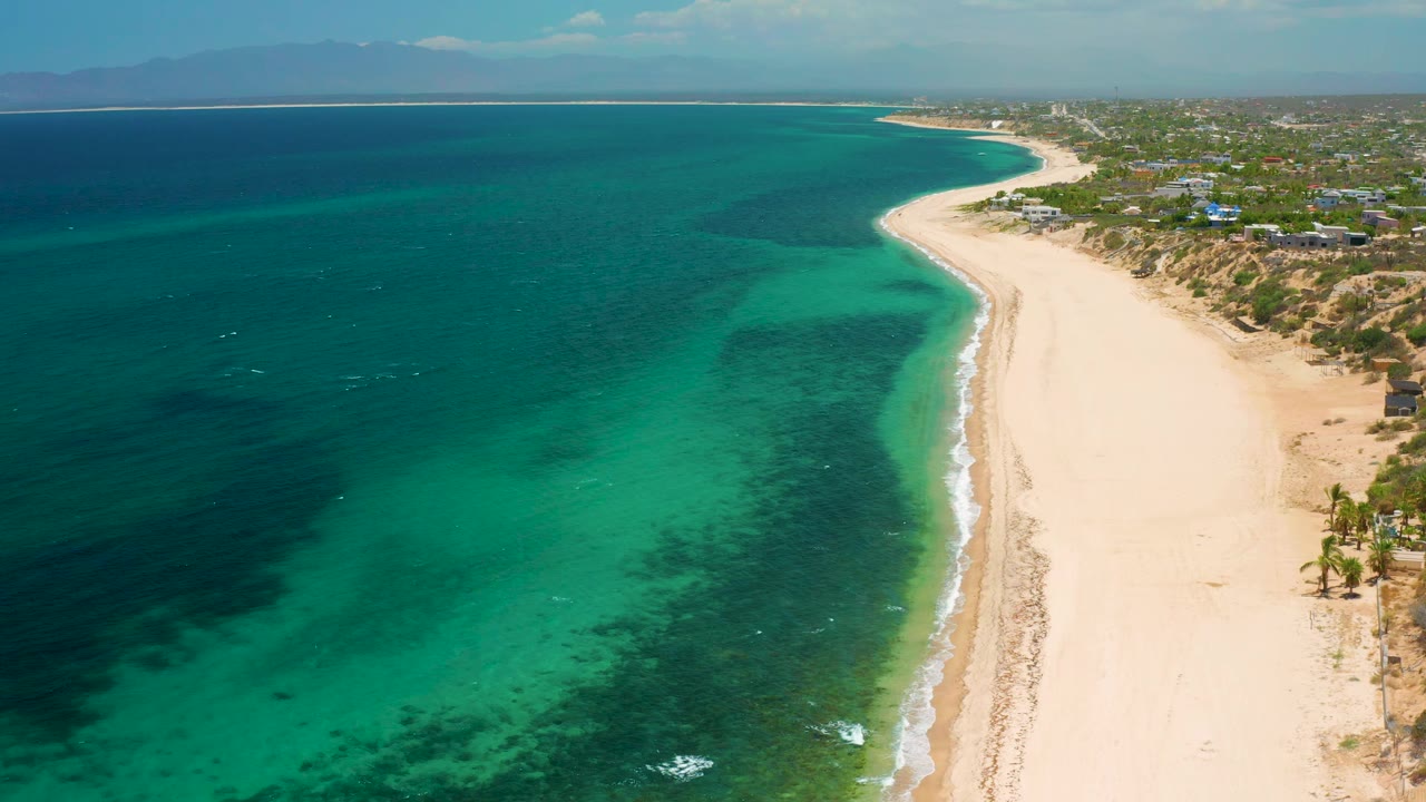Aerial view over El Sargento La Ventana, Mexico, with the beach curving into the distance, clear green water in the foreground, deeper blue water offshore, and no visible people.