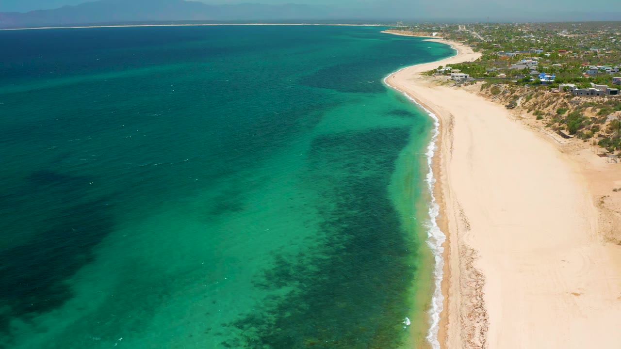 High aerial view in El Sargento La Ventana with shallow turquoise water near shore, darker water offshore, a long empty sand strip, and no visible people.