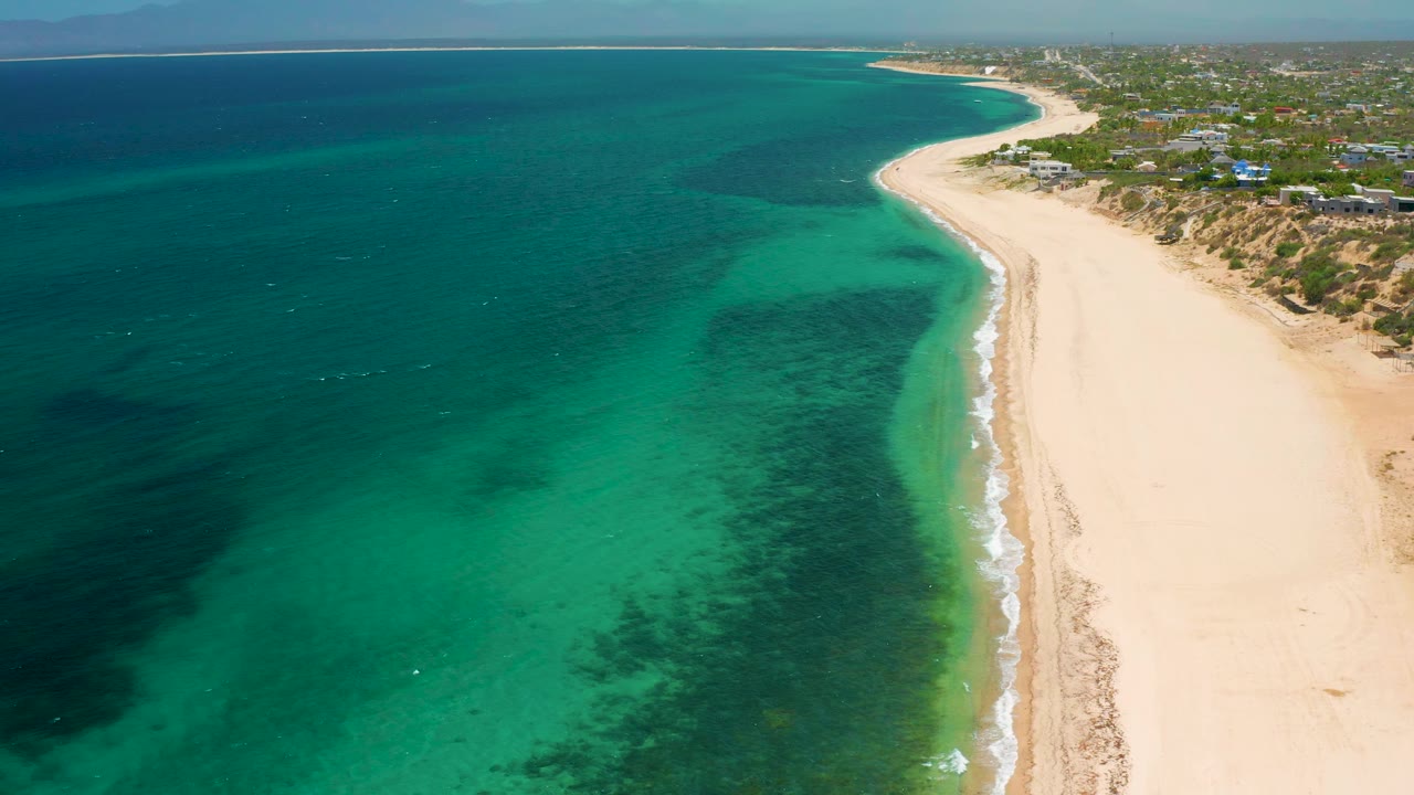 Drone-height frame over El Sargento La Ventana with turquoise shallows, darker underwater patches, pale sand, dune vegetation, distant buildings, and no visible people.
