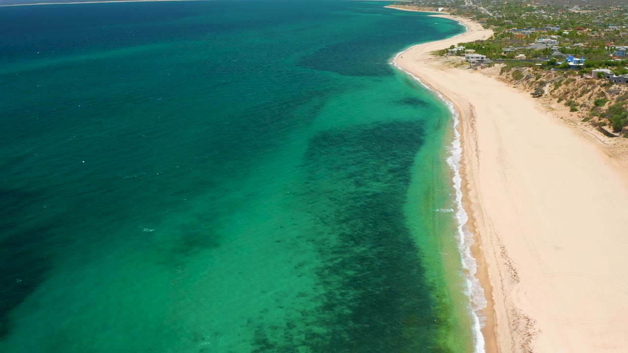 A high drone view in El Sargento La Ventana, Mexico, with the beach curving toward the upper right, darker water offshore, reef-like patches in the shallows, and no visible people.