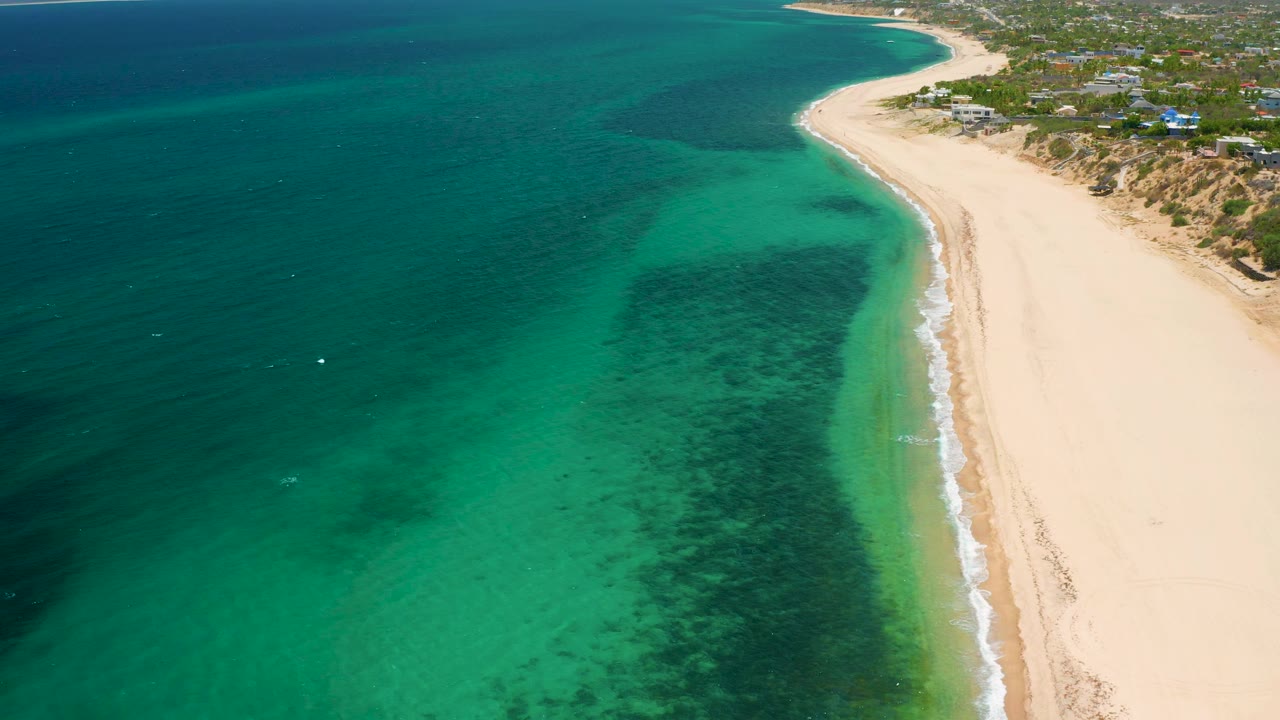 Aerial frame over El Sargento La Ventana, Mexico, looking along an empty sand beach, bright aqua shallows, darker offshore water, small surf, and no visible people.