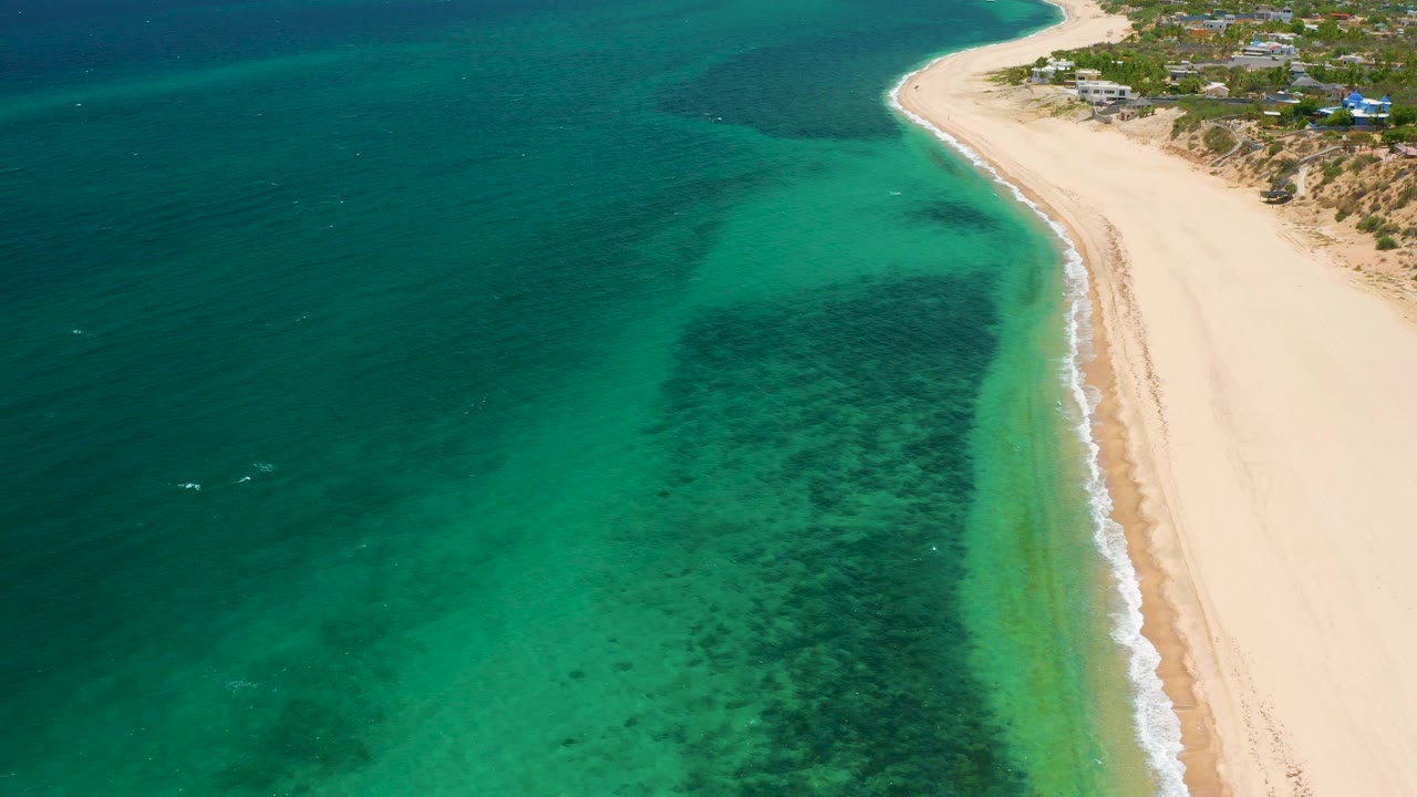 Aerial view in El Sargento La Ventana, Mexico, showing turquoise shallows, a dark underwater patch, pale sand along the right edge, and no visible people.