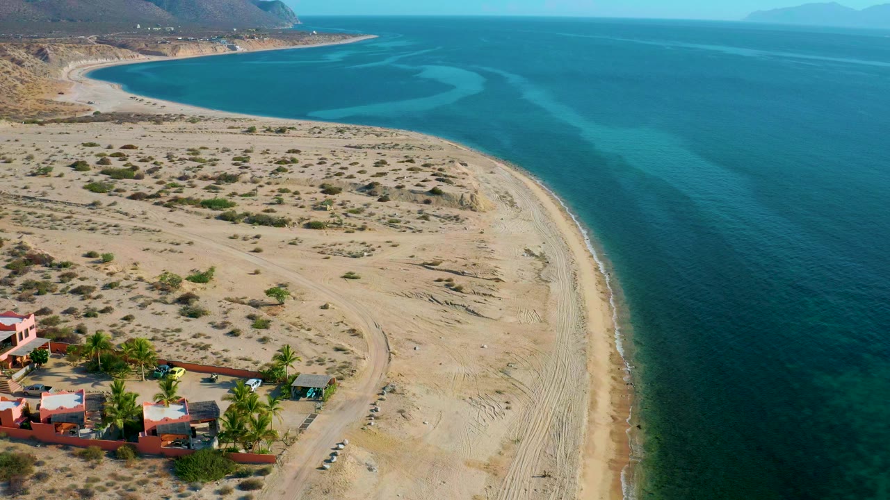 Aerial frame of El Sargento La Ventana showing sandy beach curving beside clear blue water, desert scrub inland, low coastal buildings at lower left, distant mountains, and no visible people.