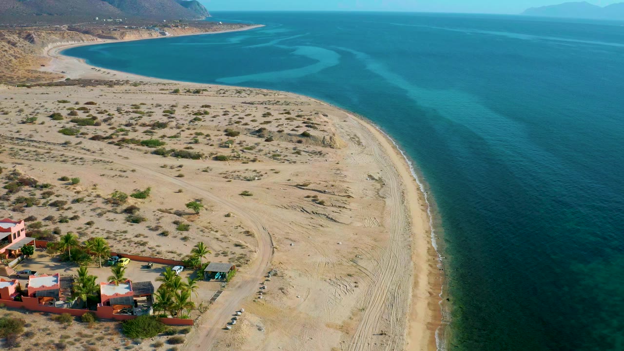 High coastal view with curved beach, teal water, scrubland, and small buildings.