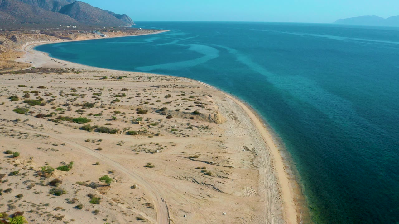 Aerial frame from El Sargento La Ventana, Mexico showing pale sand, scrubby foreground, a curved shoreline, blue-green ocean, mountain background, and no visible people.