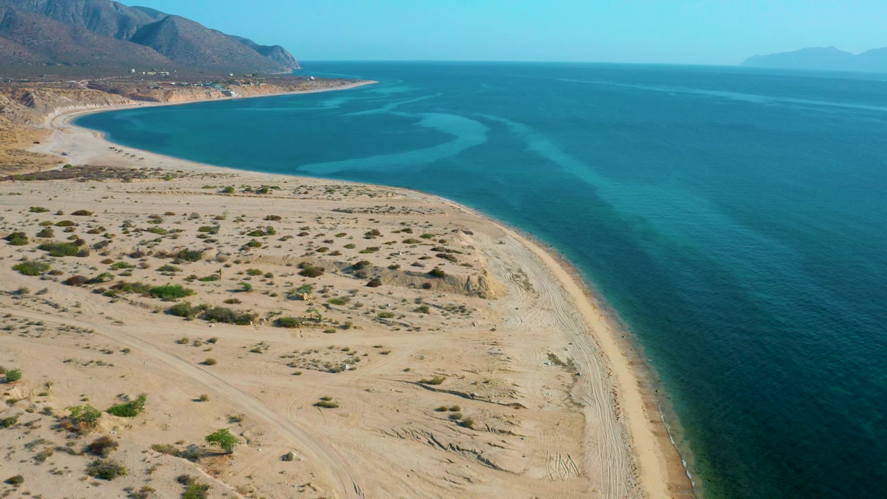 El Sargento La Ventana coastline seen from above, with the beach edge running down the right side of the sand, open blue ocean filling the right half, distant land on the horizon, and no visible people.