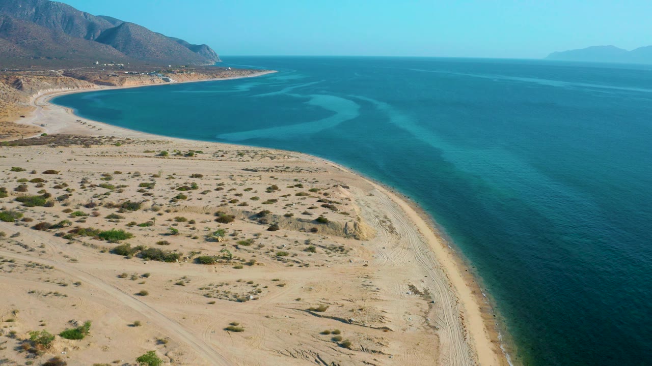 High aerial frame over El Sargento La Ventana, Mexico showing textured sand and scrub in the foreground, a narrow beach strip, calm blue-green water, mountains to the left, and no visible people.