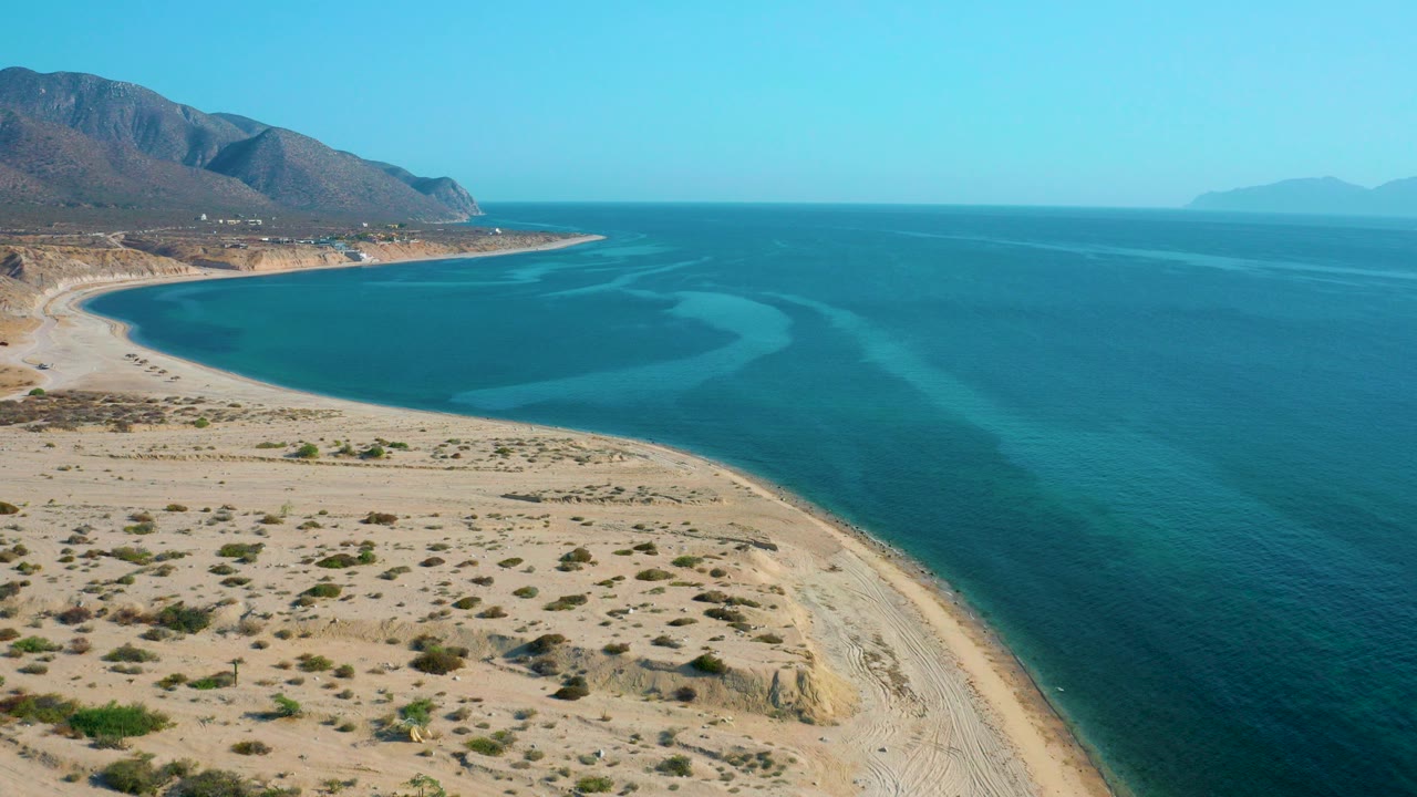 A high aerial frame over El Sargento La Ventana, Mexico shows pale sand and sparse desert shrubs below, blue-green water curving into the bay, brown hills along the left coast, and no visible people.