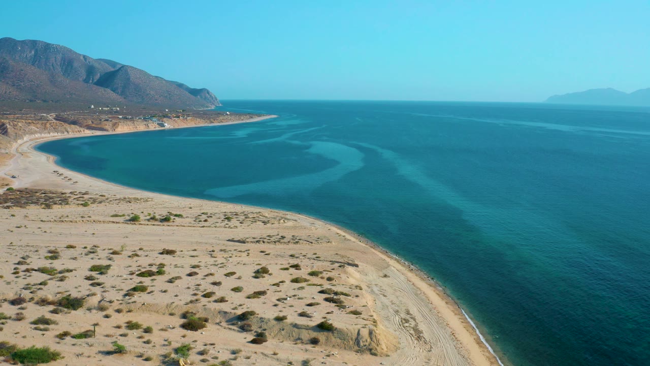 At 0:02, the aerial view over El Sargento La Ventana includes more open water to the right, a pale beach running diagonally through the lower frame, brown mountains on the left, and no visible people.