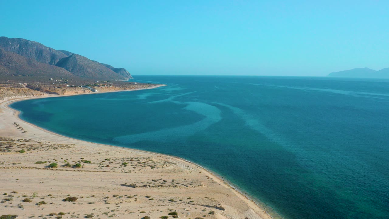 Aerial frame over El Sargento La Ventana, Mexico showing a curved sandy shoreline, blue bay, left-side mountains, distant horizon haze, and no visible people.