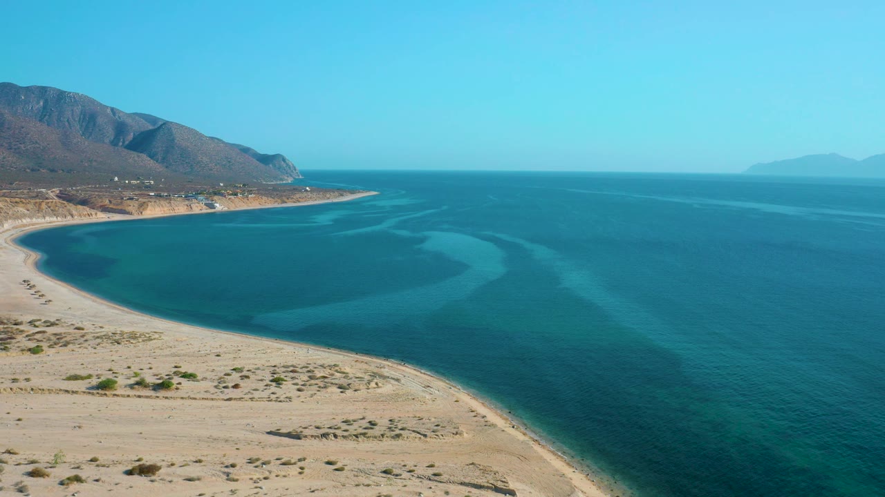 Aerial frame over El Sargento La Ventana, Mexico showing the sandy desert shoreline curving around a blue bay, with small coastal structures near the mountain base and no visible people.