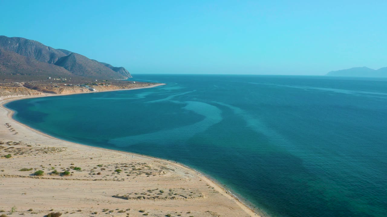 Wide aerial coast with sandy foreground, blue water, and distant mountains