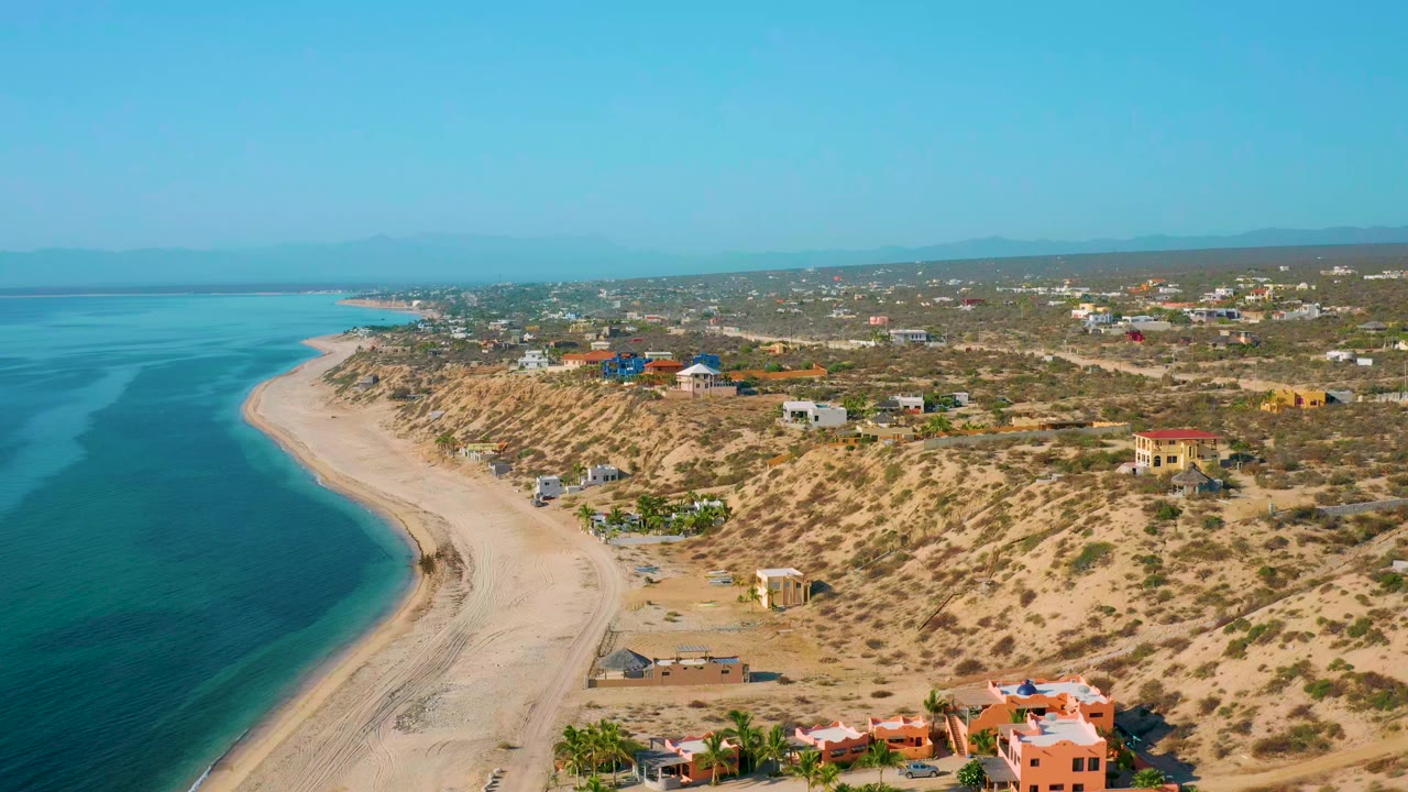 Aerial view in El Sargento La Ventana, Mexico, with turquoise water curving along pale sand, scrub dunes, small buildings, and no visible people.