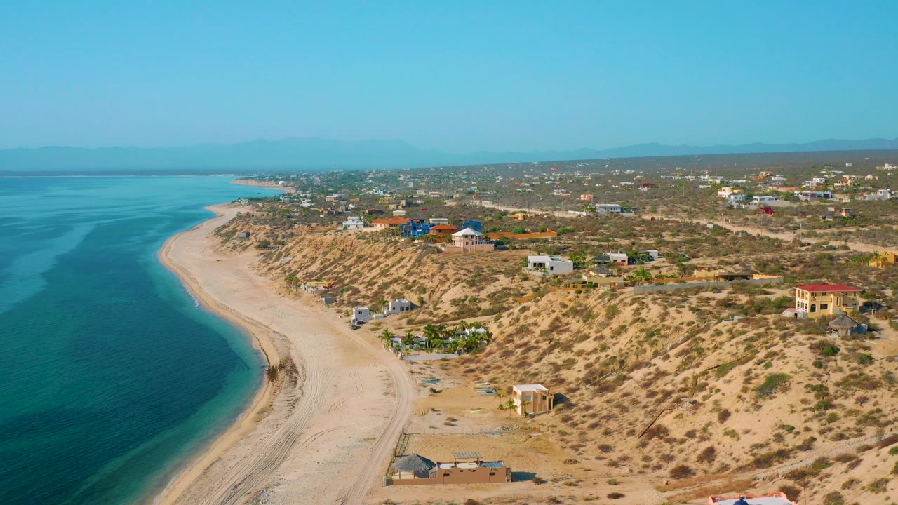 Opening aerial frame over El Sargento La Ventana, Mexico, showing blue-green water on the left, a curved sand beach, scrub dunes, scattered houses, distant hills, and no visible people.